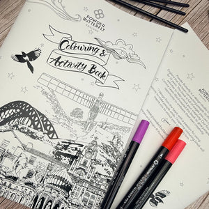 A Powdered Butterfly 'Colouring & Activity Book' spread showing black-and-white illustrated pages, with three colored markers placed on top on a wood surface.
