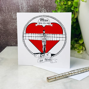 Card with a red heart and Angel of the North illustration, featuring text 'Mam, you are my Angel of the North'. on a table next to a plant with a silver pen