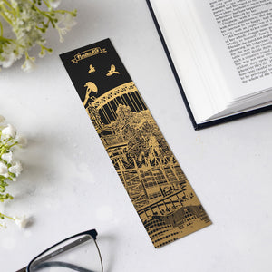A long rectangular black and gold bookmark with an illustrated Newcastle city architectural scene and the title 'Newcastle' at the top, placed on a light surface with an open book, glasses, and small white flowers around it.