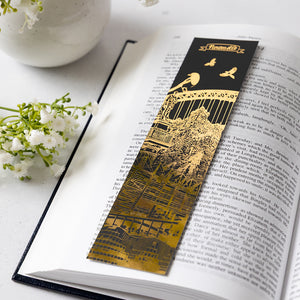A long rectangular black and gold bookmark with an illustrated Newcastle city architectural scene and the title 'Newcastle' at the top, placed on an open book with small white flowers around it.