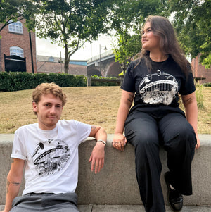 Two models sitting on concrete steps outdoors. The person on the left wears a white graphic Newcastle themed T-shirt and gray pants, while the person on the right wears a black Newcastle themed graphic T-shirt and black pants.