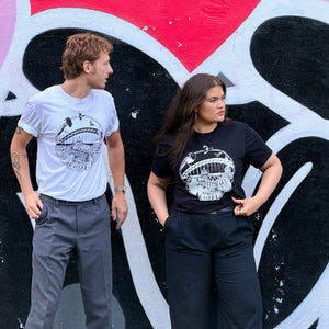 Two models standing side-by-side in front of a colourful Mul Draws mural, wearing matching black and white Newcastle themed graphic t-shirts with a variety of Newcastle landmarks and magpies.