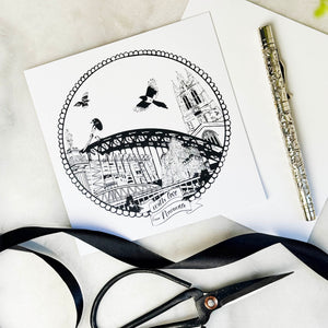 Black and white illustration of a cityscape on a card with scissors and ribbon on a light surface.