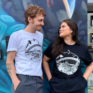 Two models standing side-by-side in front of a blue mural, wearing matching black and white Newcastle themed graphic t-shirts with a variety of Newcastle landmarks and magpies.
