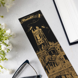 Decorative bookmark with gold illustration of bumble bees, a cathedral and 'Manchester' text on a white surface with flowers and glasses.