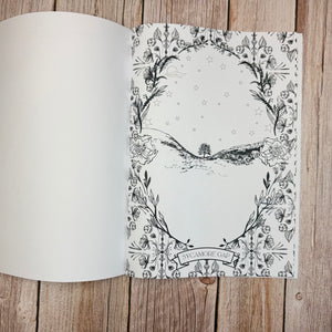 An open colouring book laid flat on a rustic wooden surface, showing a blank left page and an illustrated right page with a black-and-white botanical border, stars, and sycamore gap. The words 'SYCAMORE GAP' appear at the bottom of the illustrated page.