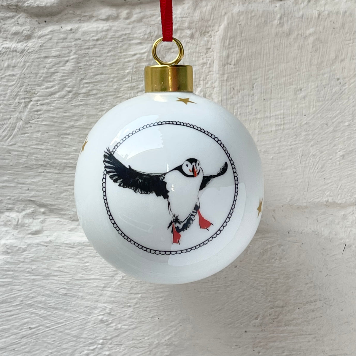 A white Christmas ornament featuring a black and gray puffin bird design with orange feet, hanging from a red ribbon.