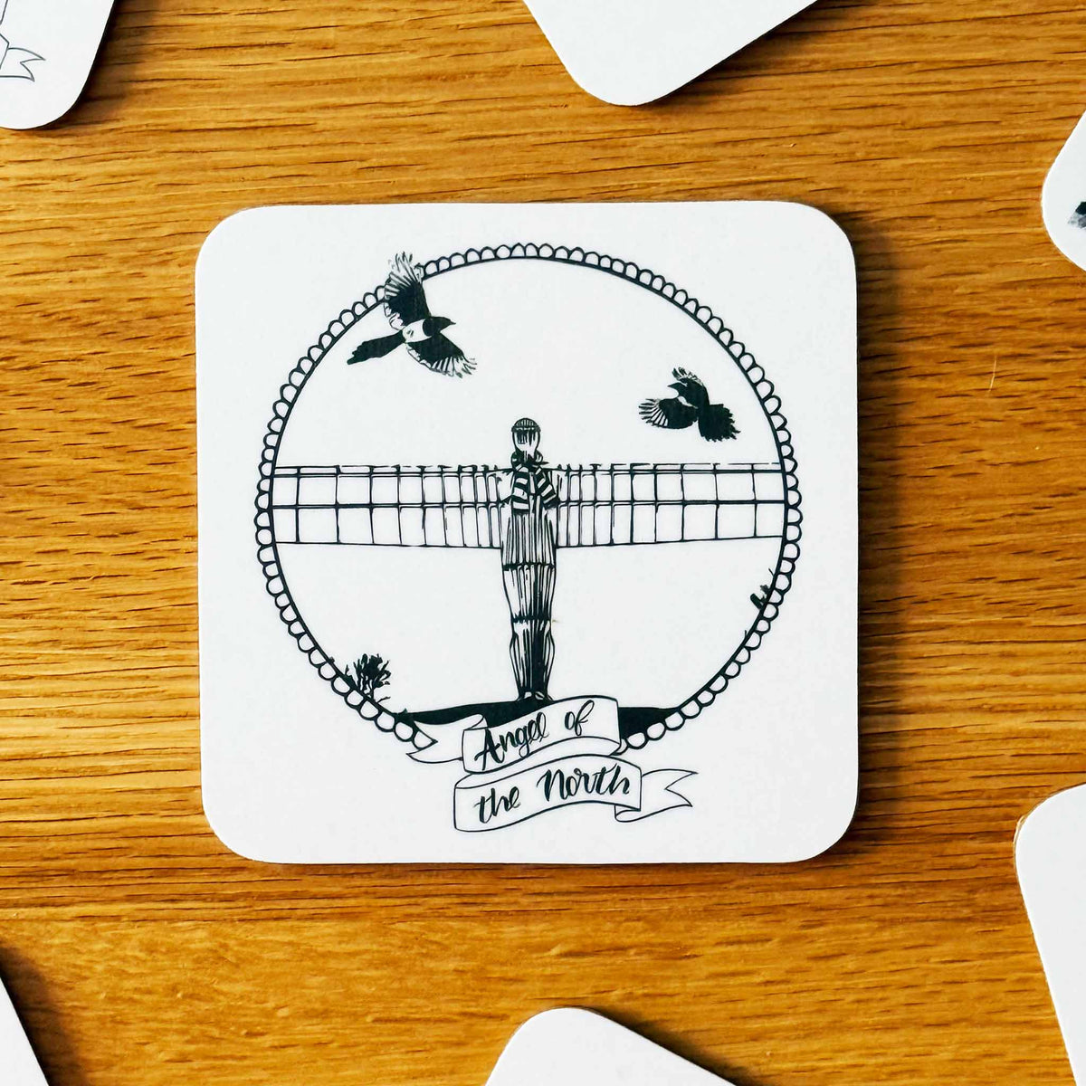 A square white coaster with rounded corners featuring a black line art illustration of the Angel of the North statue, two magpies, and the text 'Angel of the North' on a wooden tabletop.