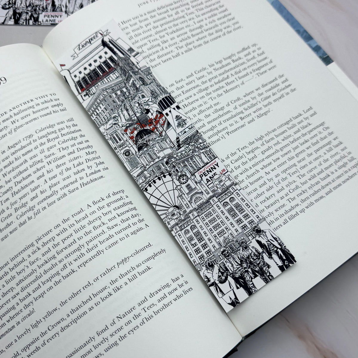 A black and white illustrated bookmark featuring Liverpool landmarks and sculptures with small red accents, shown resting on an open book page.