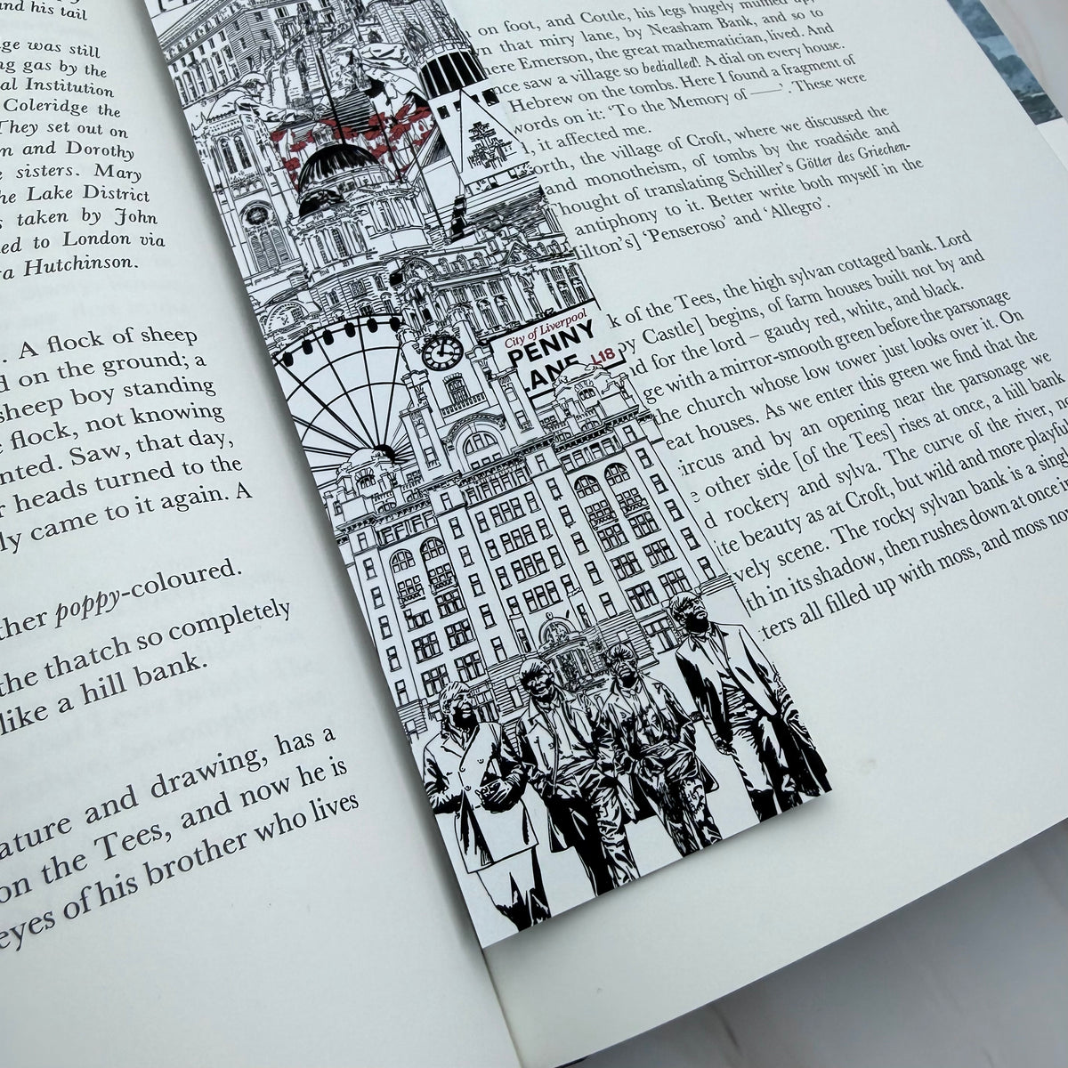 A black and white illustrated bookmark featuring Liverpool landmarks and sculptures with small red accents, shown resting on an open book page. Featuring The Beatles and Penny Lane road sign. 