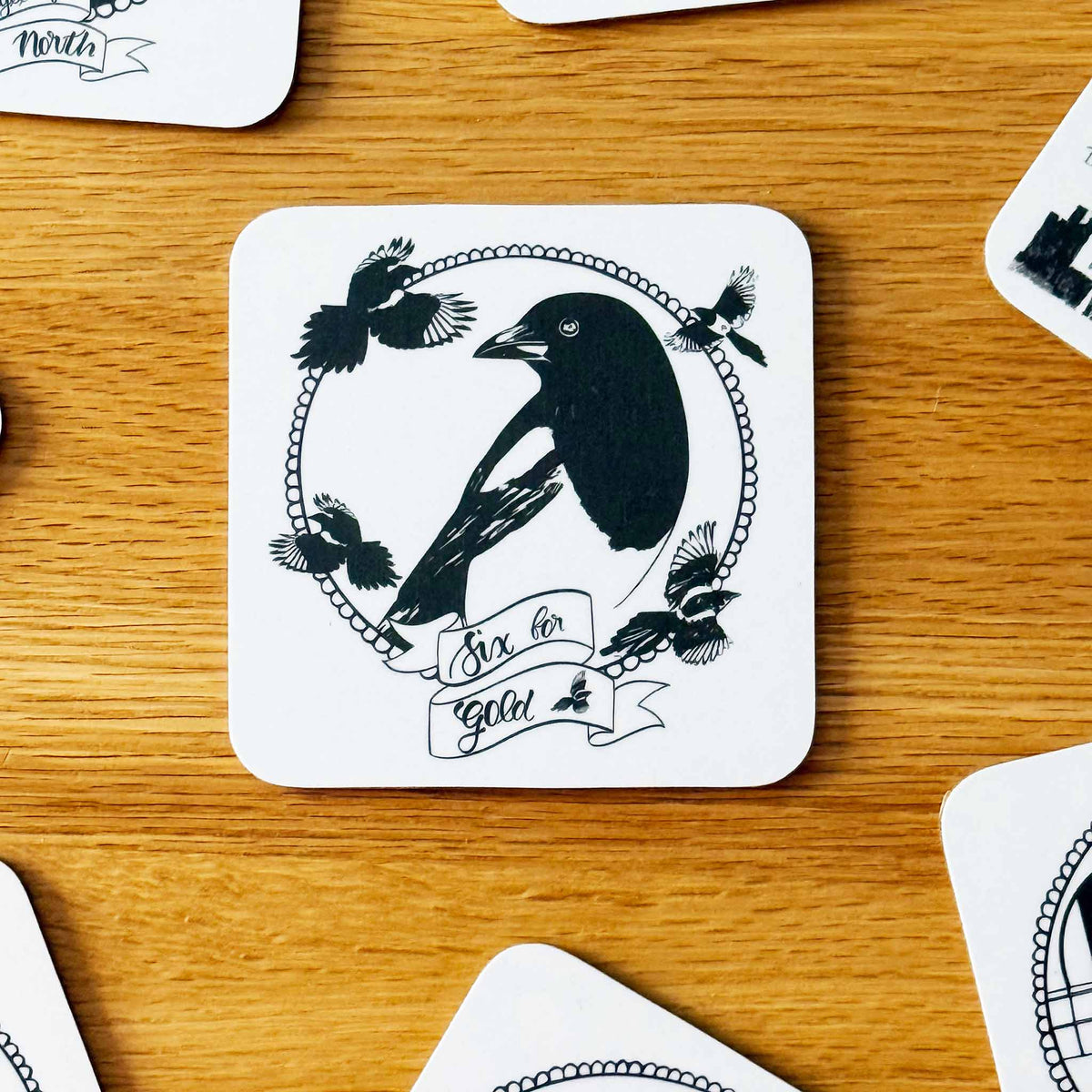 A square white coaster with rounded corners featuring a black illustration of 6 Magpies inside a circular frame with banners reading 'Six for Gold', placed on a wooden tabletop. Partially visible coasters surround it on the table.