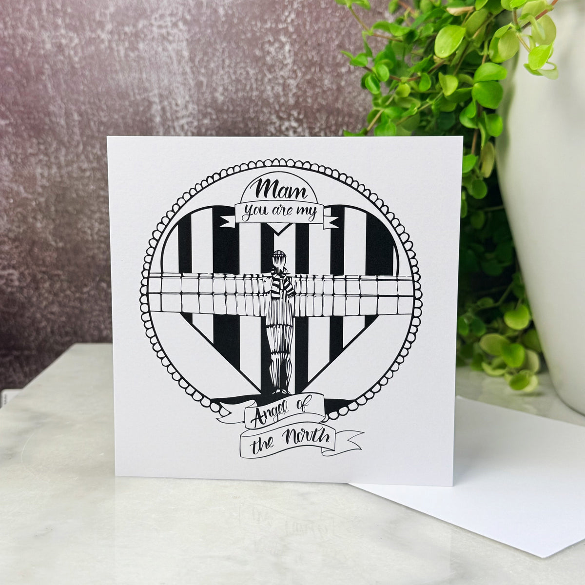 Black and white angel of the north card with the text 'mam you are my angel of the north' on a marble table with a green plant behind it 
