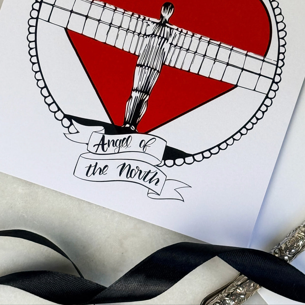 Card with a red heart and Angel of the North design, featuring the text 'Angel of the North'.  with black ribbon and a silver pen on a marble table. 