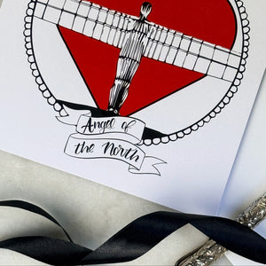 Card with a red heart and Angel of the North design, featuring the text 'Angel of the North'.  with black ribbon and a silver pen on a marble table. 