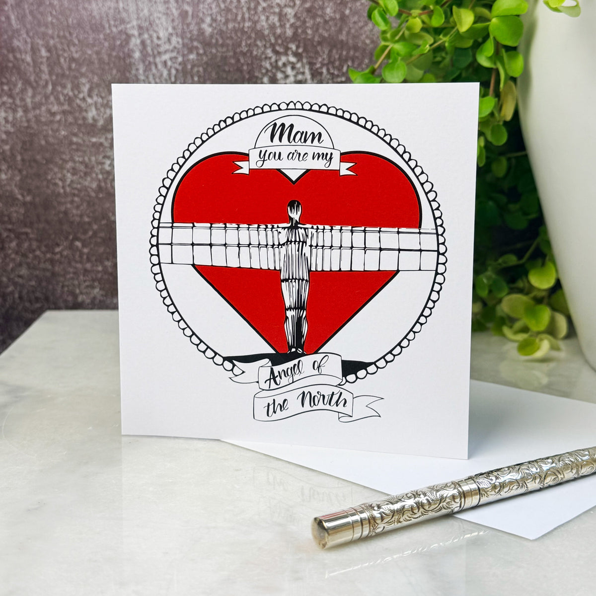 Card with a red heart and Angel of the North illustration, featuring text 'Mam, you are my Angel of the North'. on a table next to a plant with a silver pen