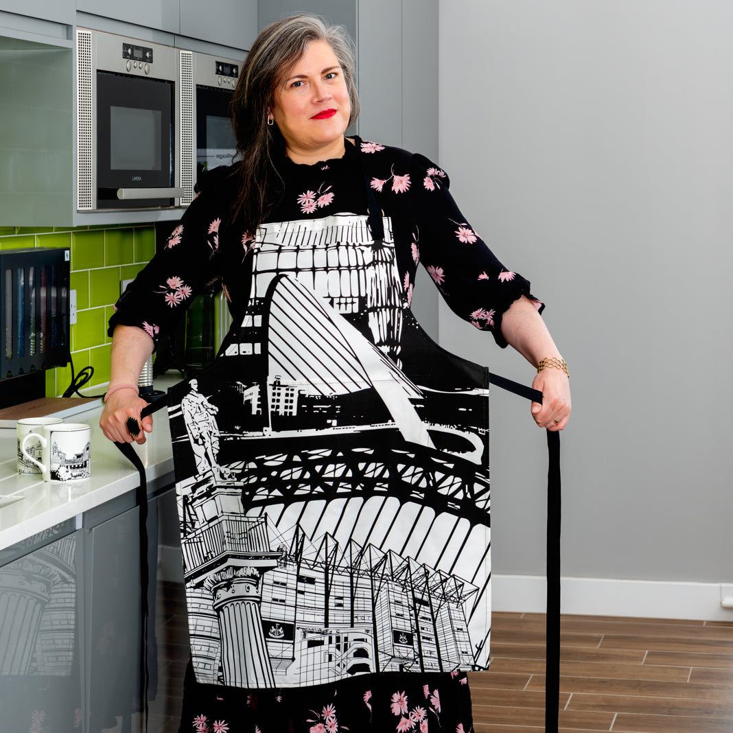 Woman standing in a kitchen holding her apron string to tie them. The apron is a bold design of Newcastle Landmarks in black and white.