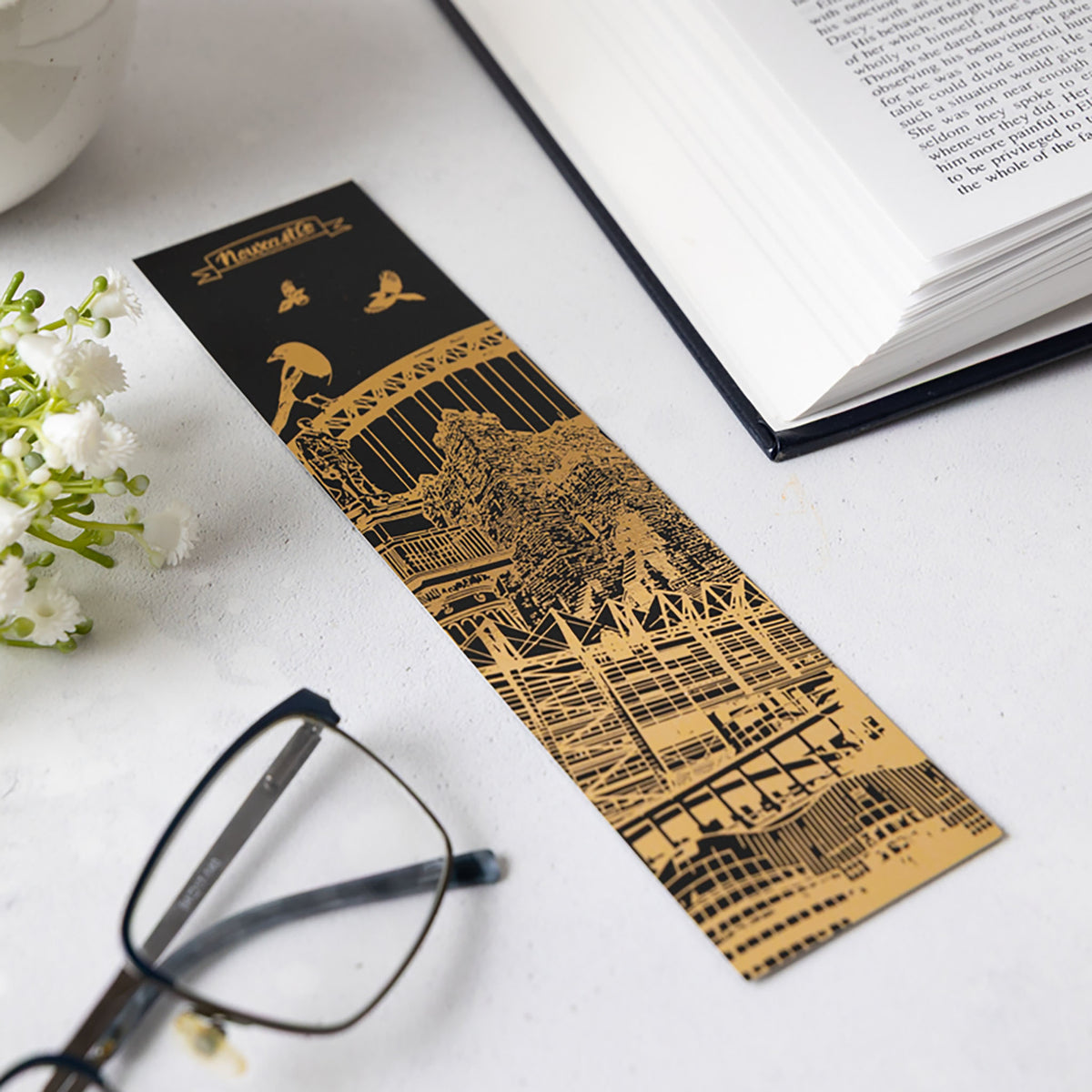 A long rectangular black and gold bookmark with an illustrated Newcastle city architectural scene and the title 'Newcastle' at the top, placed on a light surface with an open book, glasses, and small white flowers next to it.
