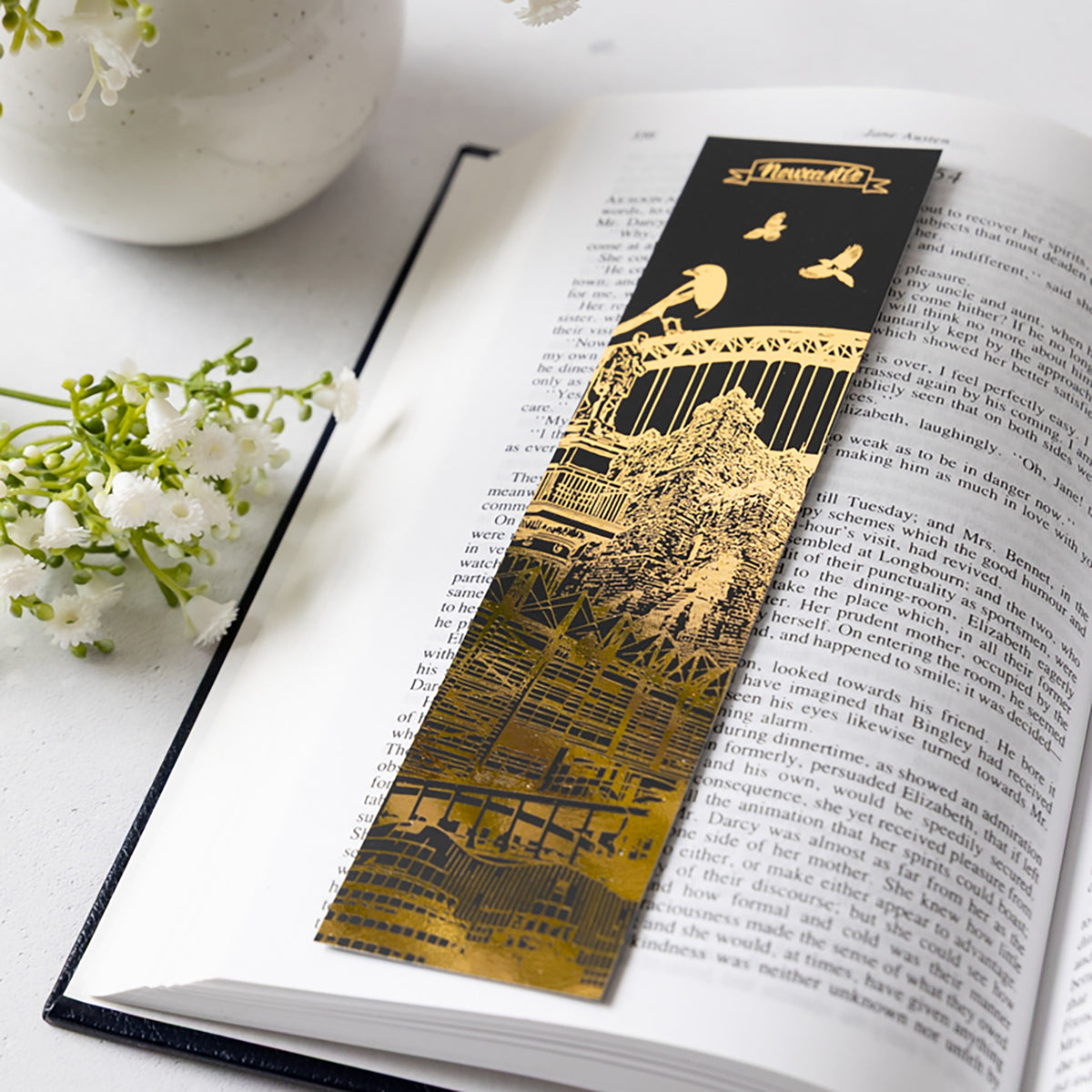 A long rectangular black and gold bookmark with an illustrated Newcastle city architectural scene and the title 'Newcastle' at the top, placed on an open book with small white flowers around it.