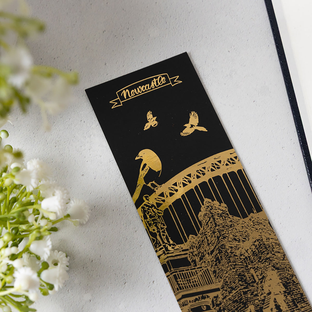 A black rectangular bookmark or card featuring gold foil illustrations of a magpie, Tyne Bridge, Newcastle Castle, Grey's Monument and the word 'Newcastle' at the top, photographed on a light textured surface with white flowers at the side.