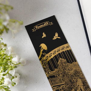 A black rectangular bookmark or card featuring gold foil illustrations of a magpie, Tyne Bridge, Newcastle Castle, Grey's Monument and the word 'Newcastle' at the top, photographed on a light textured surface with white flowers at the side.