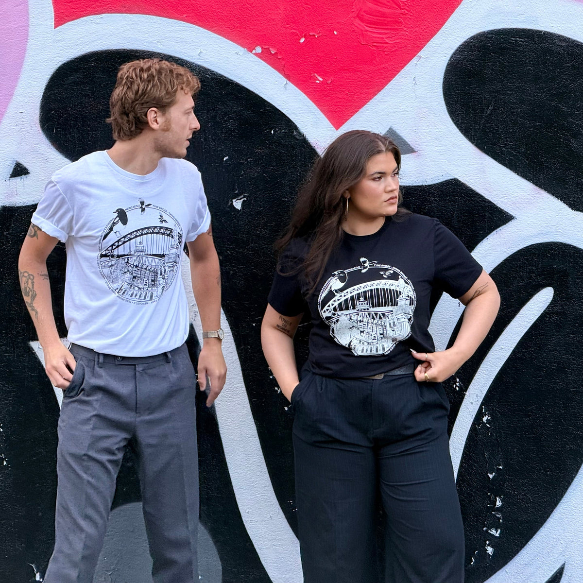 Two models standing side-by-side in front of a colourful Mul Draws mural, wearing matching black and white Newcastle themed graphic t-shirts with a variety of Newcastle landmarks and magpies.
