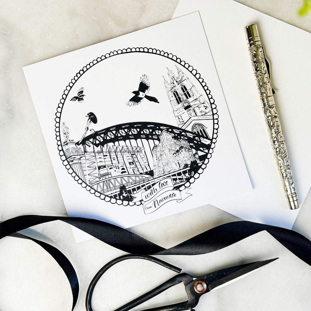 Black and white illustration of a cityscape on a card with scissors and ribbon on a light surface.