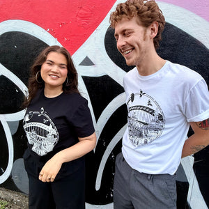 Two models standing in front of a colourful Mul Draws graffiti wall, wearing matching black and white Newcastle themed graphic t-shirts with a variety of Newcastle landmarks and magpies.