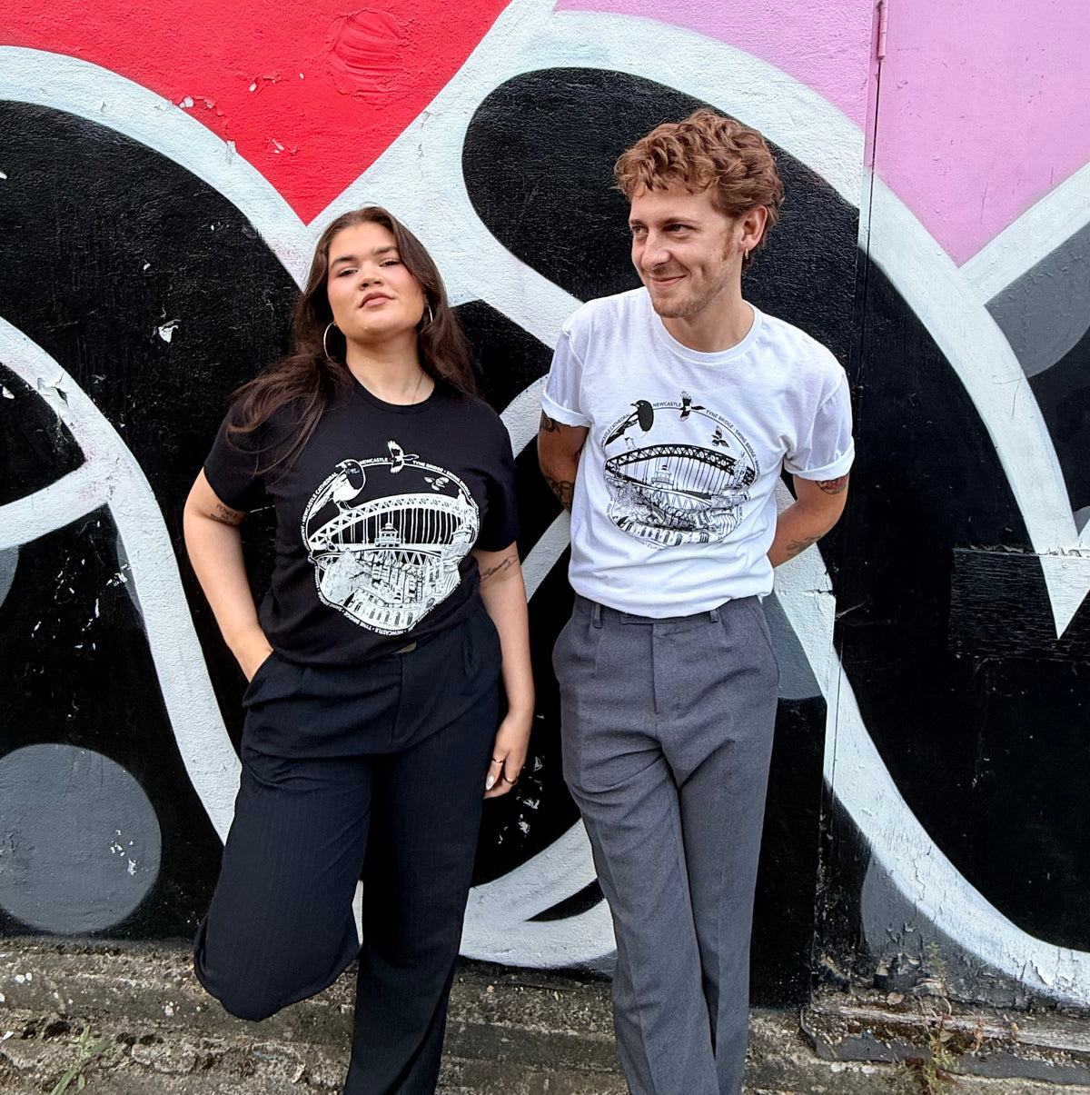 Two models standing in front of a colourful Mul Draws graffiti wall, wearing matching black and white Newcastle themed graphic t-shirts with a variety of Newcastle landmarks and magpies.