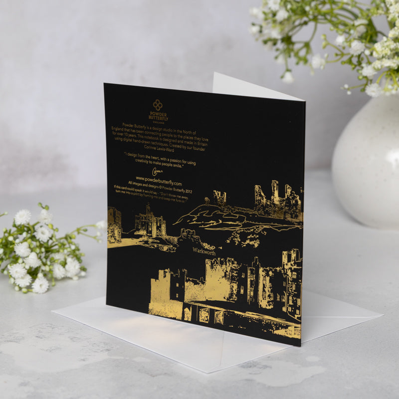Greeting card with gold foil Northumberland Castles design on a black background, placed on a white surface with flowers in the background.