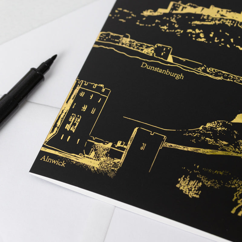 Black greetings card with gold foil artwork of Northumberland castles on a white surface with a pen.