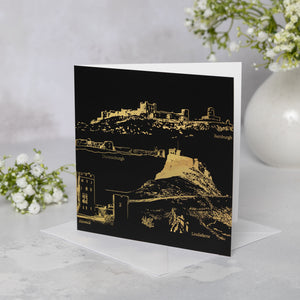 Greeting card with gold Northumberland castles illustration on a black background, placed on a white surface with flowers.