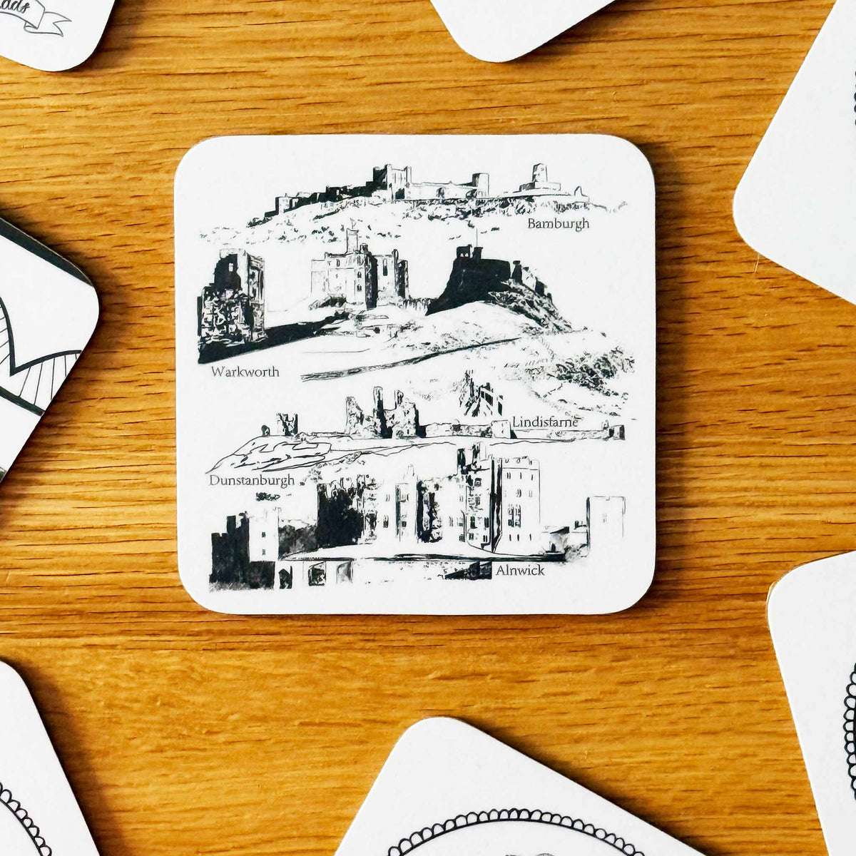 Square coaster with rounded edges on a wooden coffee table. Coaster is white with black illustration of Northumberland castles. 
