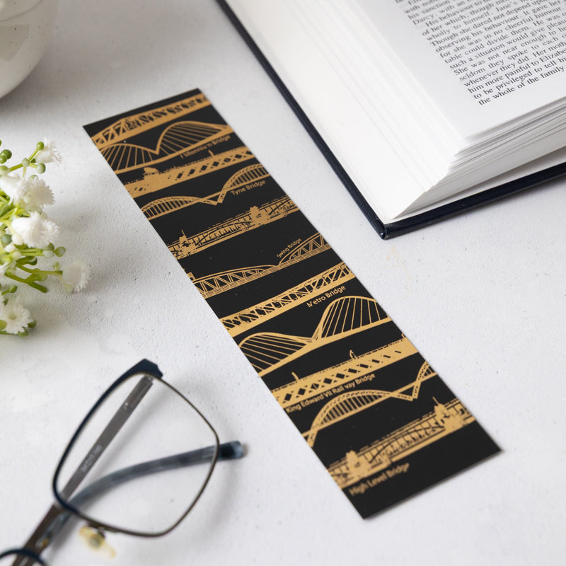 Bridges bookmark laid on a white surface next to white flowers an open book and a pair of black framed glasses.