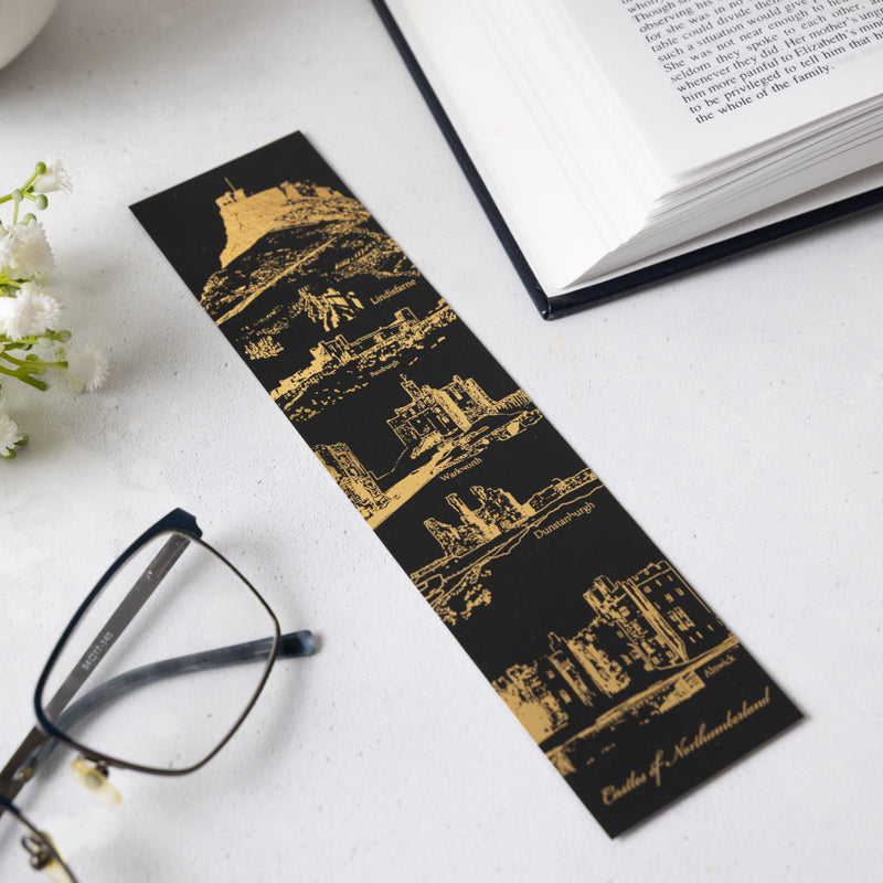 Black soft touch bookmark with gold foil castle design on a white background with an open book in the corner. 