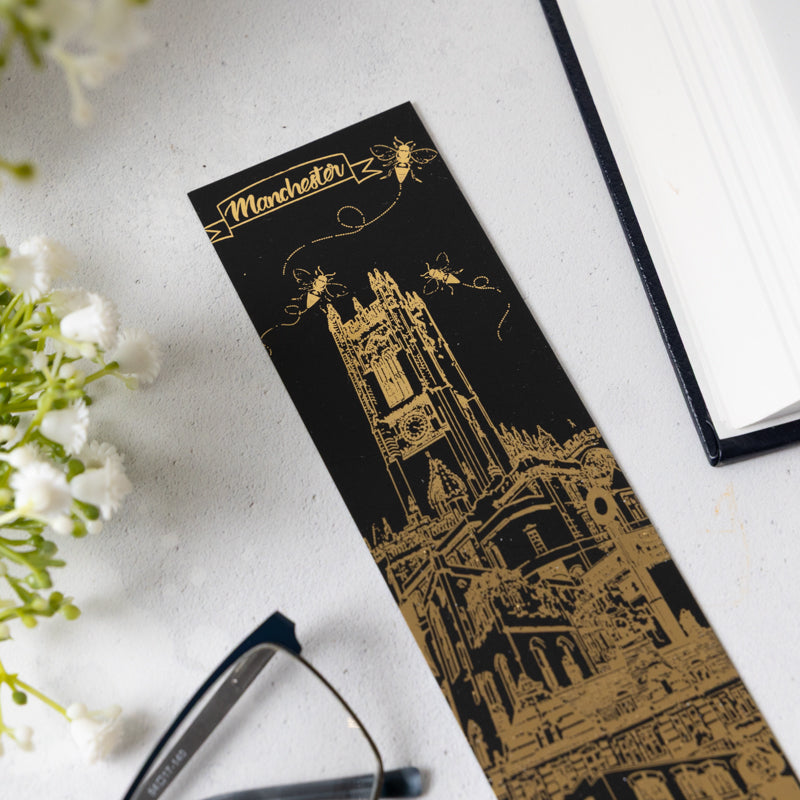 Decorative bookmark with gold illustration of bumble bees, a cathedral and 'Manchester' text on a white surface with flowers and glasses.