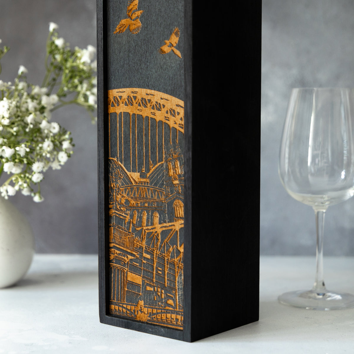 Decorative wine box with laser-engraved Newcastle architectural design on a grey background with a wine glass and flowers.
