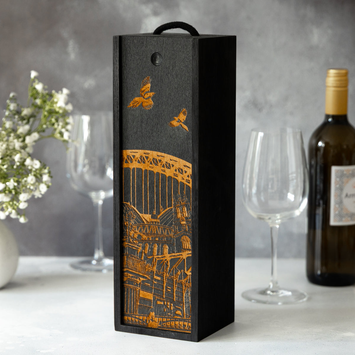 Decorative wine box with laser-engraved Newcastle architectural design on a grey background on a table with a wine bottle, wine glasses and white flowers.