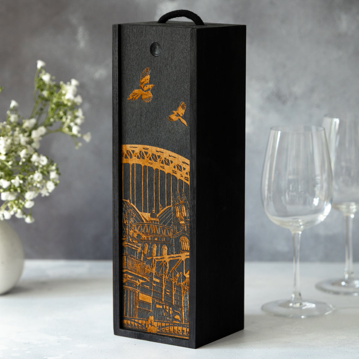 Decorative wine box with laser-engraved Newcastle architectural design on a grey background on a table with wine glasses and white flowers.