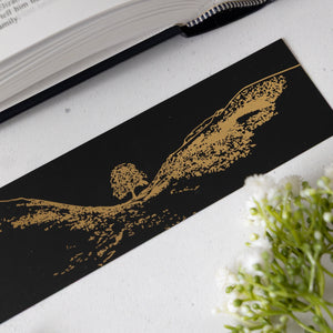 Black bookmark with gold Sycamore Gap tree design on a white surface with an open book and greenery. This is a quality Northumberland and Sycamore Gap Gift