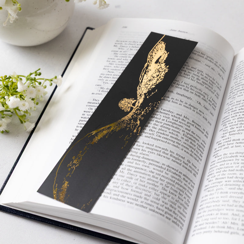 Black and gold bookmark with illustration of Sycamore Gap tree on an open book with flowers in the background