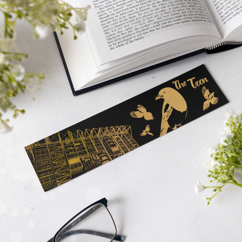 Black and gold bookmark with illustrations on a white surface with an open book and glasses.