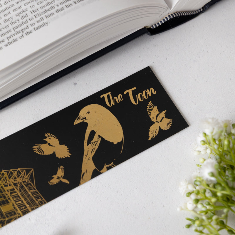 Black bookmark with gold illustrations of birds and text on a white surface with an open book.