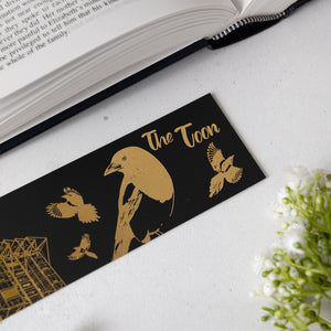 Black bookmark with gold illustrations of birds and text on a white surface with an open book.