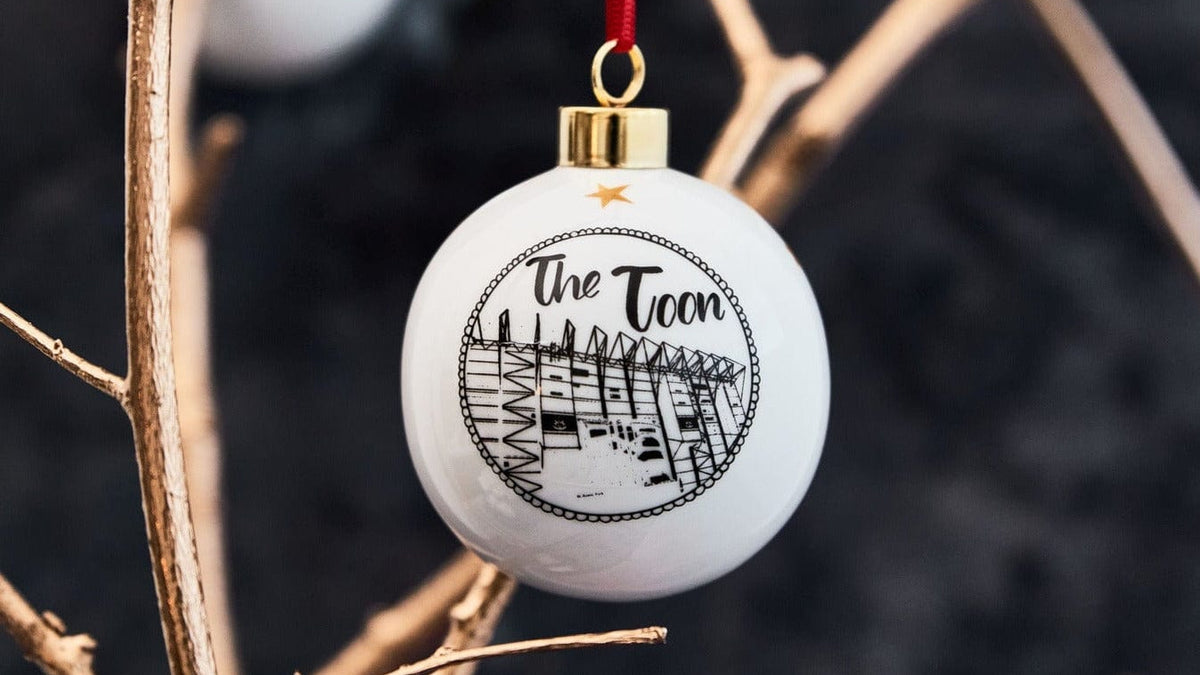 White ceramic holiday ornament featuring st james' park in a black and white illustration. design comes with a red ribbon and gold trim 