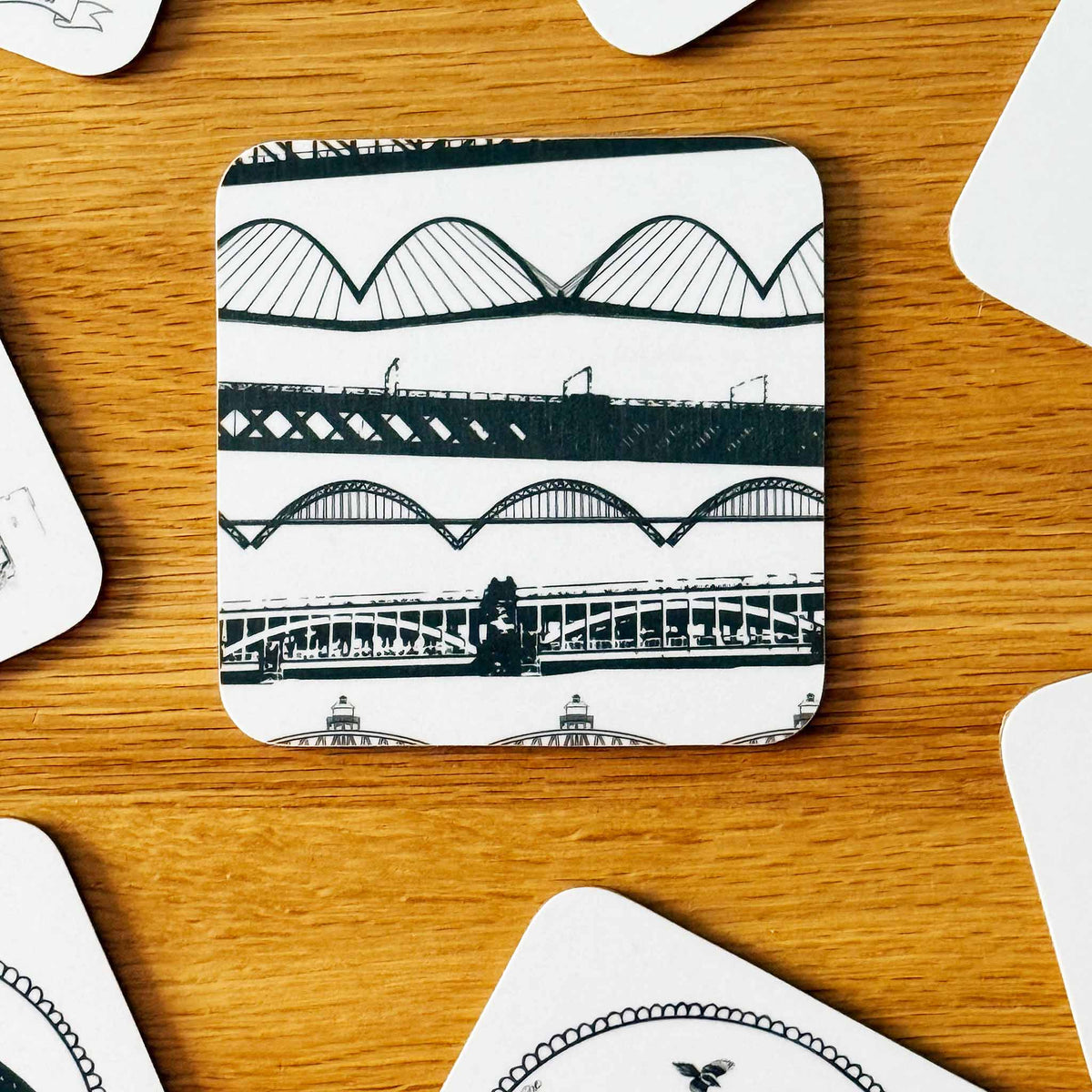 A square coaster with rounded corners featuring a black and white architectural bridge pattern of Newcastle's iconic Swing Bridge, Millennium Bridge, Tyne Bridge and High Level Bridge. The coaster is placed on a wooden tabletop with other coasters partially visible around it.