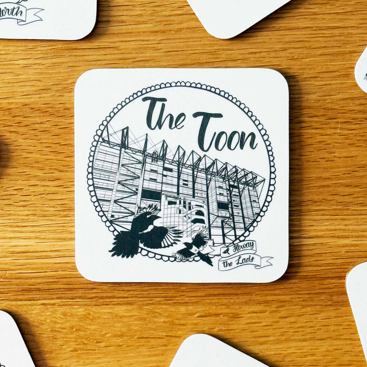 A square white coaster with rounded corners featuring a black line art illustration of St James' Park stadium and the text 'The Toon', additional text 'Howay the Lads' is in the bottom right corner, with two magpies next to it on the left-hand side. The coaster is placed on a wooden table with other coasters partially visible around it.