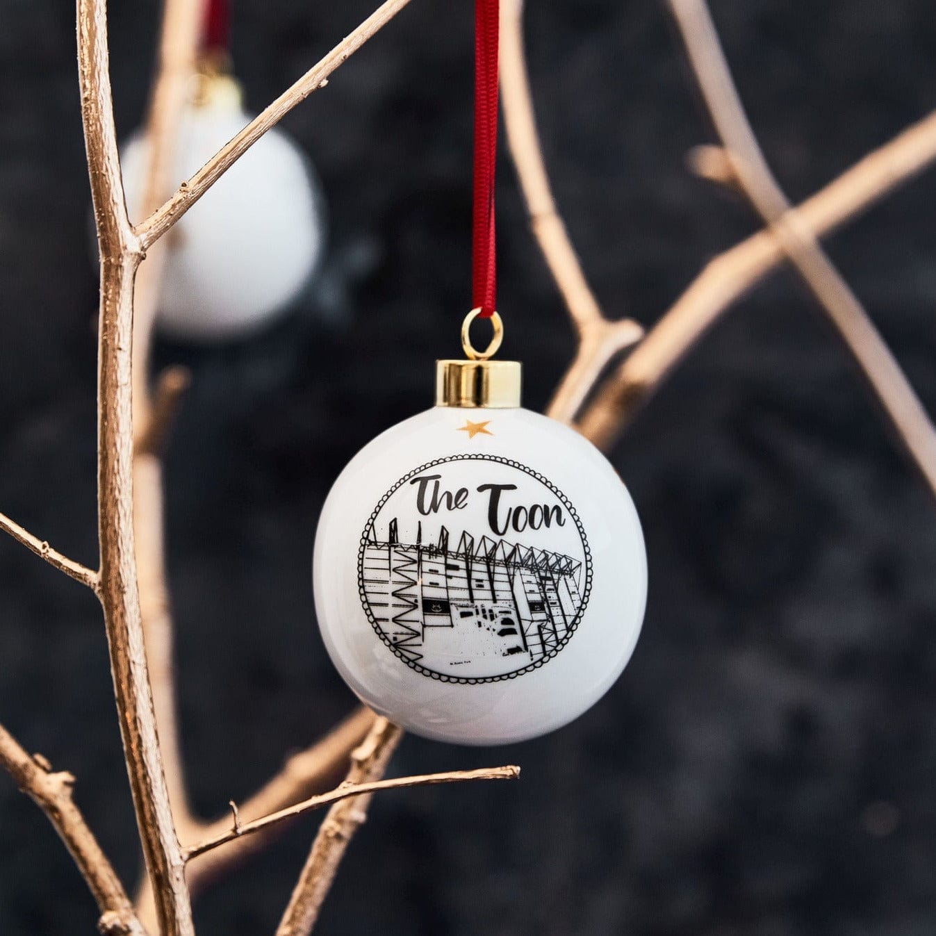 White ceramic holiday ornament featuring st james' park in a black and white illustration. design comes with a red ribbon and gold trim 