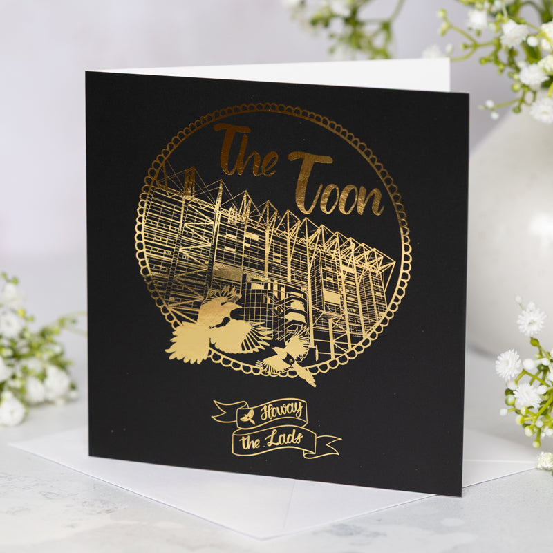 Black card with gold foil St James' Park design and text 'The Toon' on a white background with flowers.