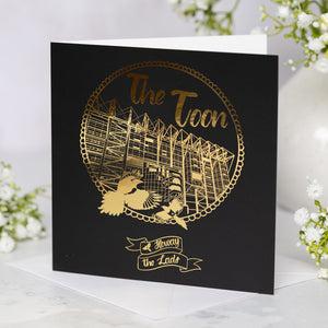 Black card with gold foil St James' Park design and text 'The Toon' on a white background with flowers.