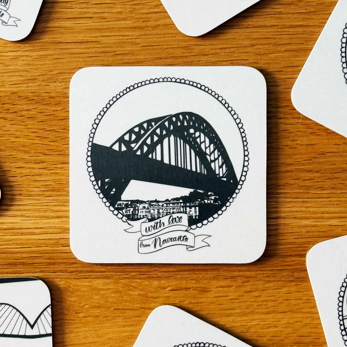 A square white coaster with rounded corners featuring a black line art illustration of the Tyne Bridge in Newcastle, surrounded by a decorative border and a banner reading 'with love from Newcastle'. The coaster is placed on a wooden tabletop with other coasters partially visible around it.